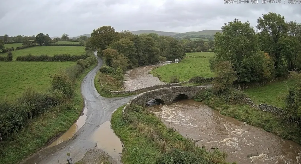 Flood: Generative AI report. Timed capture. Generative AI reports swollen brown water near the top of the banks, the road wet but passable, level about 1.1 m. The rate-of-rise rule warns the duty engineer; no closure yet.
